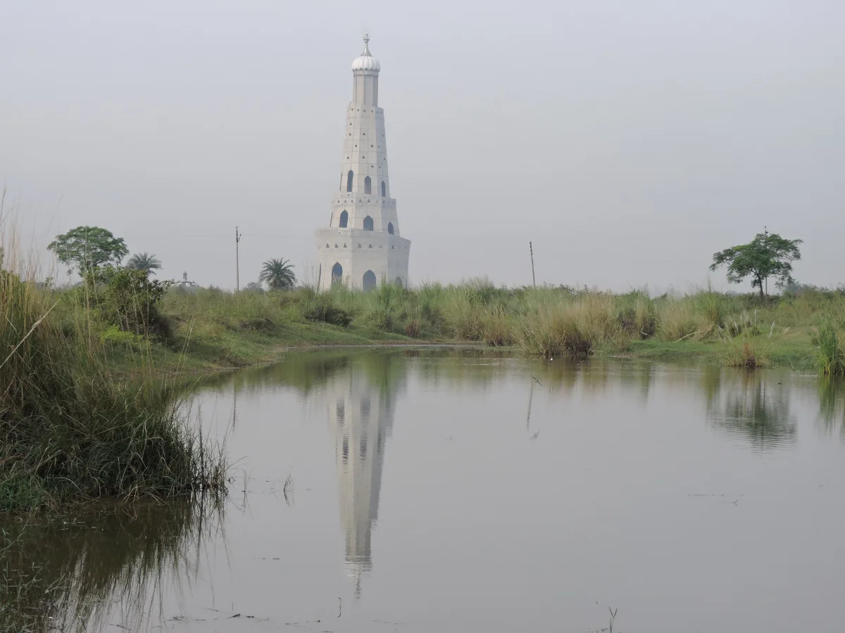 Tall white tapering Fateh Burj tower at Chappar Chiri rising above grass and trees, with its reflection in still pond water in the foreground