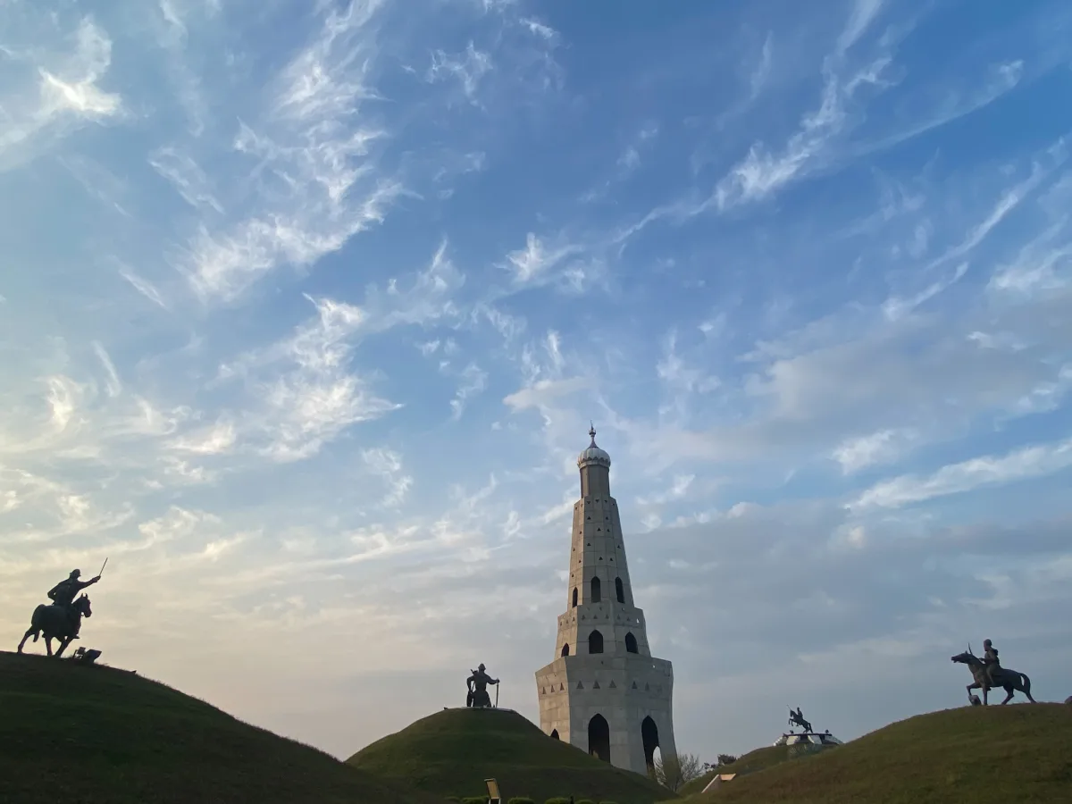 Fateh Burj tower against a blue sky with wispy clouds, with equestrian statues in silhouette on grassy mounds on either side