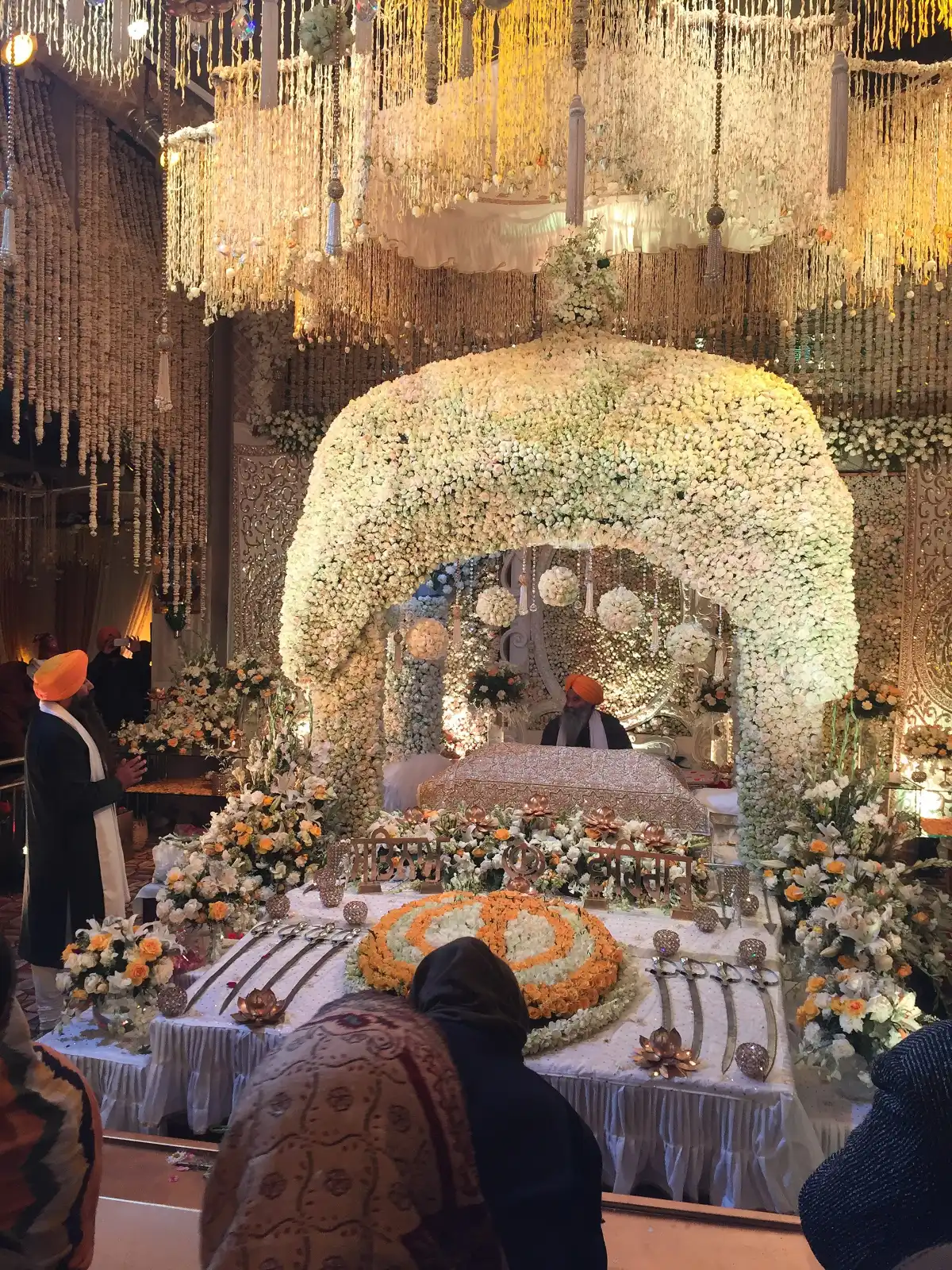 Nada Sahib Gurdwara, Panchkula: History, Timings And How To Reach 2 Guru Granth Sahib under a canopy of white and orange flowers inside Gurdwara Nada Sahib, with worshippers standing nearby