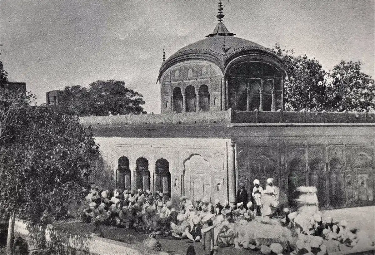 Old black and white photograph of the earlier Gurdwara Fatehgarh Sahib with a domed pavilion, arched wall and a seated crowd of devotees in the courtyard