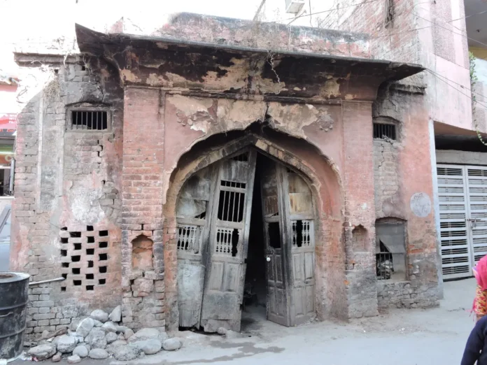 Manimajra Fort gateway, Chandigarh Weathered red brick gateway of Manimajra Fort with a pointed arch, peeling plaster and warped wooden doors, standing beside modern shops
