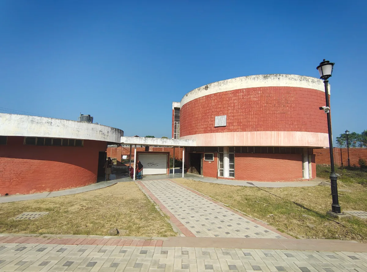 Red brick circular museum building at Sanghol with a white roof edge, a paved path across a lawn and a black lamp post under a clear blue sky
