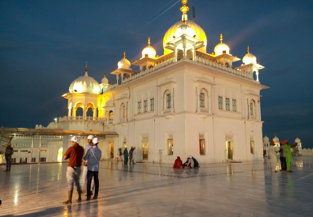 White marble Takht Sri Kesgarh Sahib lit up at dusk, with a golden dome and worshippers on the marble courtyard