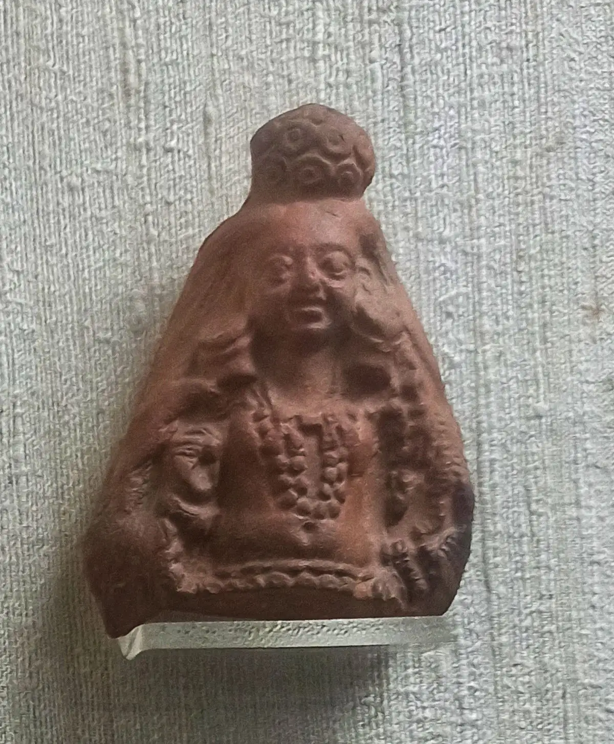 Small reddish-brown terracotta bust of a Yakshi with a topknot, layered necklaces and a smiling face, shown against a grey wall